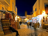 People Strolling along Cobblestone Street Past Carpet Seller at Twilight, Kairouan, Tunisia Photographic Print by Bethune Carmichael