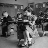 Cute Little Girl Busily at Work, Sitting in a Desk Chair in a Schoolroom, Other Pupils at Work Too Photographie par Gordon Parks