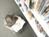 Little Boy Reading Book Beside Library Shelf Photographic Print
