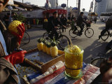 Cyclists Pass by a Street Vendor Selling Carved Pineapples Photographic Print by Justin Guariglia