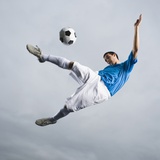 Mixed Race Teenager in Mid-Air Kicking Soccer Ball Photographic Print by Erik Isakson